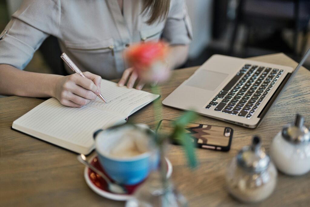 pexels-photo-1766604-1766604 Woman writing in a notebook with a laptop and coffee cup on a desk. Ideal for workspace inspiration.